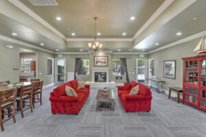 Interior Clubhouse, 2 red couches with coffee table between, contemporary gray carpeting, fireplace with landscape art above mantle, long wood dining table, red book case, exit to pool area left of fireplace.