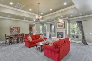 Interior Clubhouse, 2 red couches with coffee table between, contemporary gray carpeting, fireplace with landscape art above mantle, long wood dining table, red book case, exit to pool area left of fireplace, attached kitchen right of dining table.