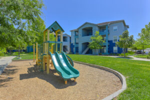 Exterior Playground, Soft ground, slides, climbing bars, benches along sidewalks nearby, residential buildings around playground.