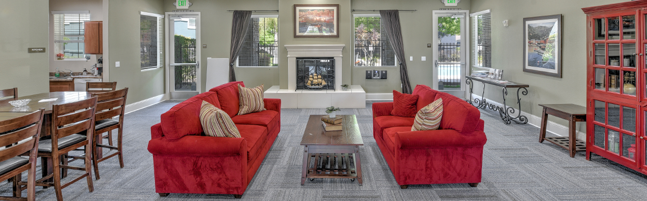 Interior Clubhouse, 2 red couches with coffee table between, contemporary gray carpeting, fireplace with landscape art above mantle, long wood dining table, red book case, exit to pool area left of fireplace.