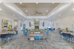 Interior Clubhouse, contemporary carpeting with hints of blue, 3 square tables with 4 seats at each, 4 blue lounge chairs, 1 gray couch, yellow accents throughout the room, landscape painting above fireplace, white walls, exit to pool area in back of room.