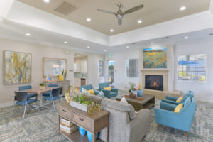Interior Clubhouse, four blue lounge chairs, square table with four chairs, attached kitchen, fireplace with landscape painting above, gray couch, rustic coffee table, exit to pool area.