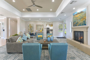 Interior Clubhouse, contemporary area rug with hints of blue, three square tables with four seats at each, four blue lounge chairs and a gray couch in center of the room, fireplace with landscape painting above it, attached kitchen, exit to pool area in back of room, white walls, rustic decor with yellow accents.