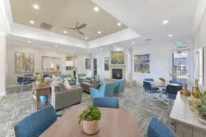 Interior clubhouse, three square tables with four blue chairs around them, four blue lounge chairs and gray couch in center of room, contemporary carpeting with blue accents, rustic decor, white walls, attached kitchen.