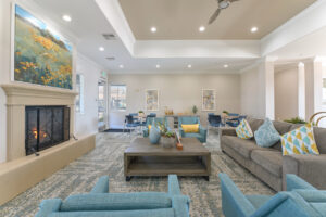 Interior Clubhouse, contemporary area rug with hints of blue, three square tables with four seats at each, four blue lounge chairs and a gray couch in center of the room, fireplace with landscape painting above it, attached kitchen, exit to pool area in back of room, white walls, rustic decor with yellow accents.