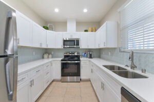 Interior clubhouse kitchen, tile floor, stainless steel appliances, dual sink, stone countertop, white cabinetry.